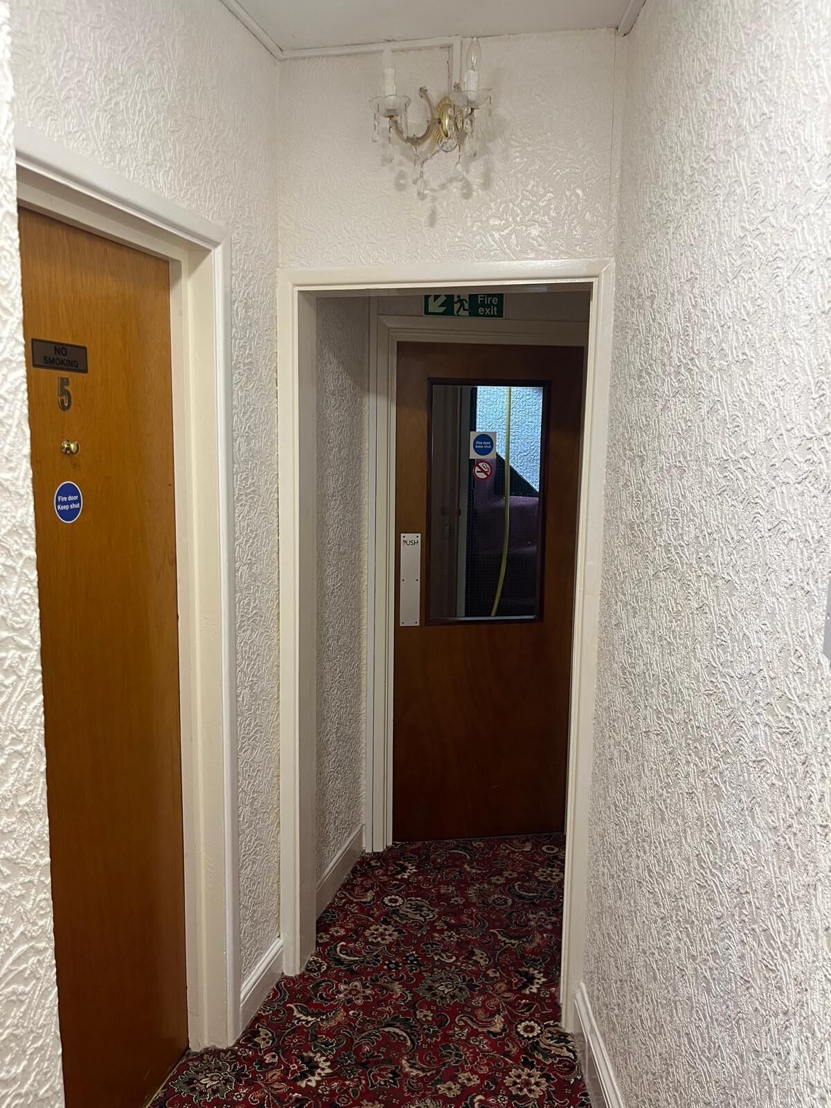 A hallway with a patterned carpet, a chandelier, a door with a 'NO SMOKING' sign, and a 'Fire exit' sign.