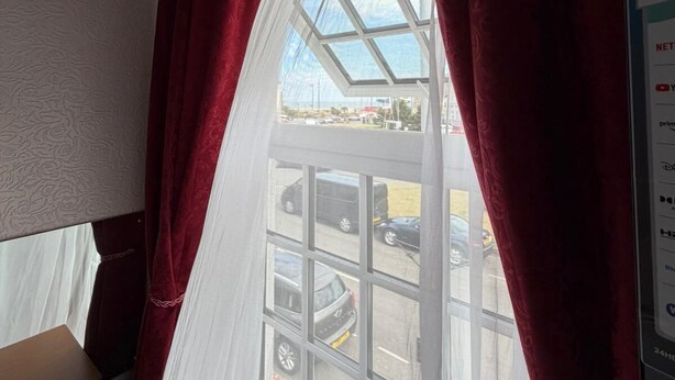 A room with a large window, red curtains, and a view of parked cars outside. - Fjaerland Hotel (Great Yarmouth)