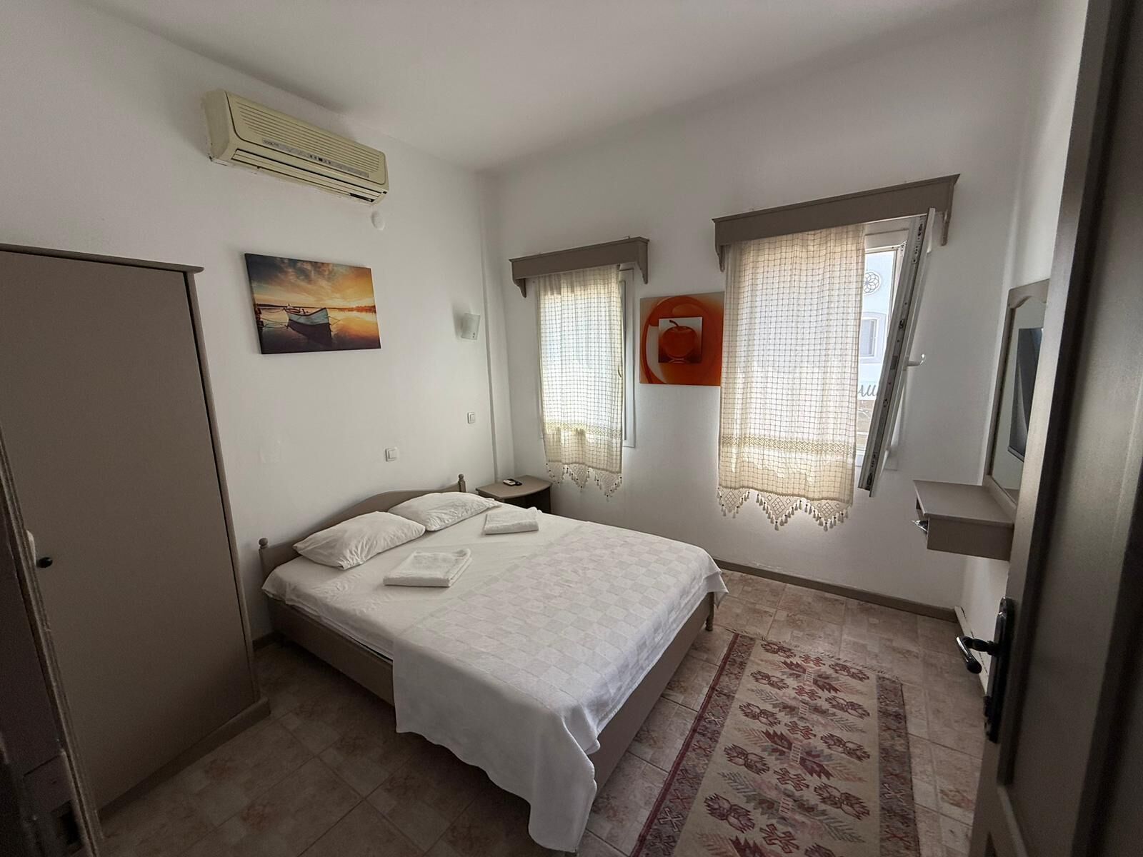 A bedroom with a bed, a nightstand, an air conditioner, a painting, and a window with curtains.