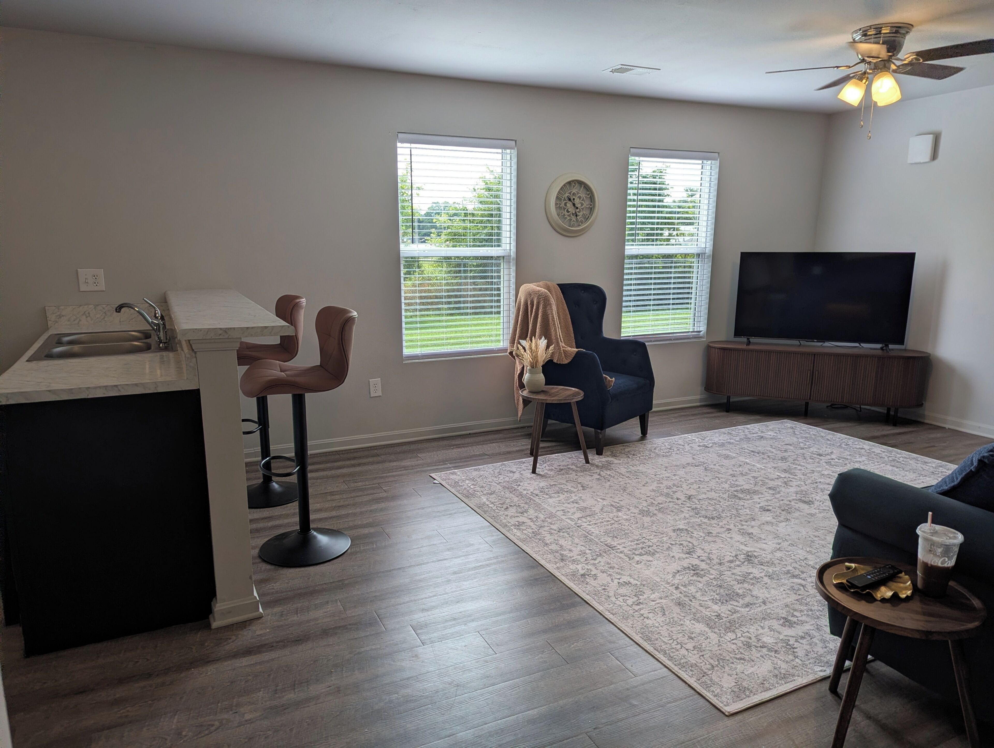 A modern living room with a kitchen area, a dark blue armchair, a sofa, a TV, and a ceiling fan.