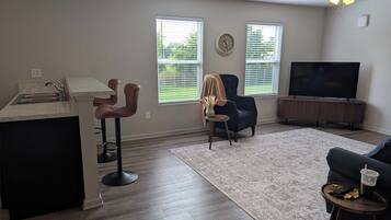 A modern living room with a kitchen area, a dark blue armchair, a sofa, a TV, and a ceiling fan.