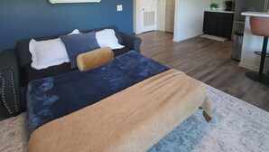 Un dormitorio con cama, mesita de noche, estante en la pared con objetos decorativos y puerta que da a un pasillo.