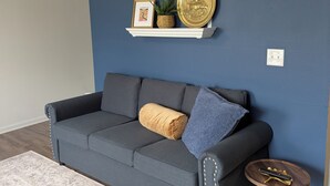 Un salón con una pared azul oscuro, un estante blanco con objetos decorativos, un sofá gris con cojines y una alfombra estampada.