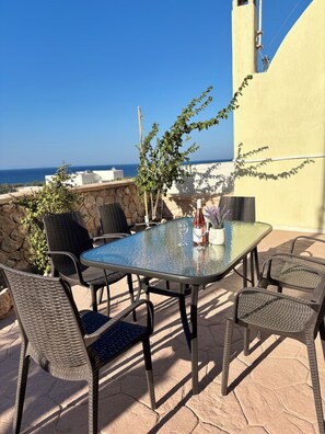Ein Tisch mit Glasplatte, Stühle, einer Flasche und einem Glas darauf, auf einer Terrasse mit Blick aufs Meer.