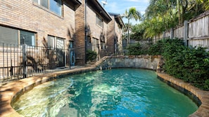 Ein Pool mit Steinrand, umgeben von einem Backsteinhaus mit Metallzaun.