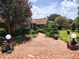 Uma passagem de tijolos leva a um edifício com telhado de azulejos, cercado por vegetação exuberante e plantas floridas.