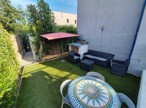 Een terras met een mozaiektafel, buitenmeubels en een rode parasol.