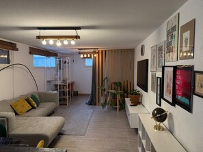 Una sala de estar moderna con un sofá, un escritorio y una estantería. Hay plantas en macetas y cuadros en las paredes.