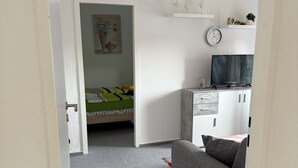 Ein Zimmer mit grauem Sofa, einem Fernseher auf einem Schrank, einem Bett mit grün-weißer Bettdecke und einer weißen Kommode mit Schubladen.