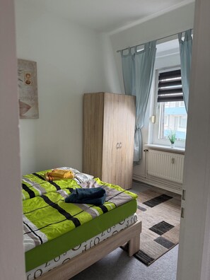 A bedroom with a bed, a wardrobe, a radiator, and a window with curtains. - 2-room apartment on the North Sea directly on the dyke (Cuxhaven)