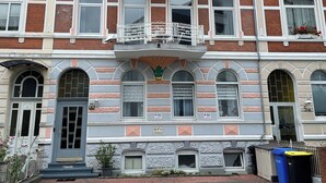 Ein mehrstöckiges Gebäude im traditionellen Baustil, mit einem zentralen Balkon mit aufwendiger Geländerarbeit, zahlreichen Fenstern mit Fensterläden und einer Ziegel-Fassade.