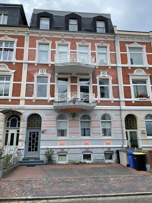 A multi-story building with a traditional architectural style, featuring a central balcony with ornate railings, multiple windows with shutters, and a brick facade. - 2-room apartment on the North Sea directly on the dyke (Cuxhaven)