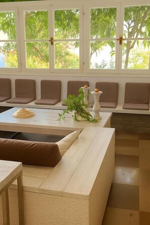 A room with a large window, a seating area with cushioned benches, a table with a plant, and a small table with a candle. - ADAMANTIA HOTEL (Samos)