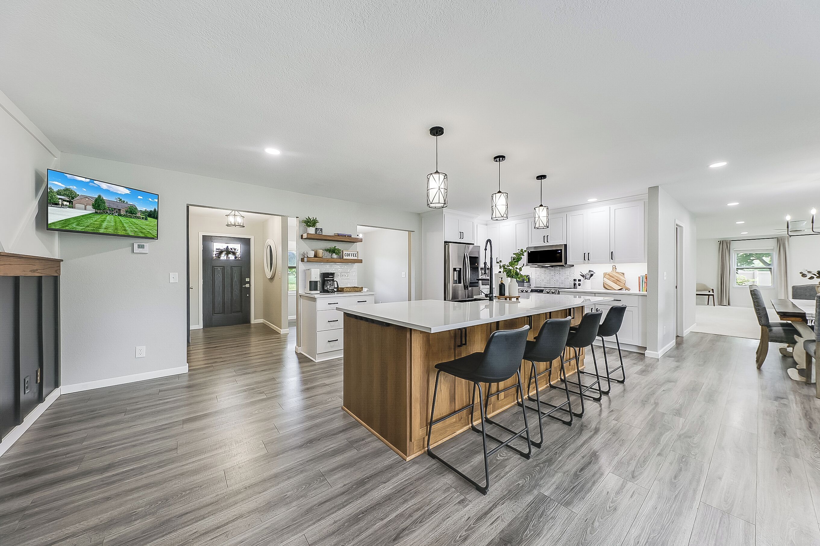 A modern kitchen with a central island, bar stools, pendant lighting, and a flat-screen TV mounted on the wall.