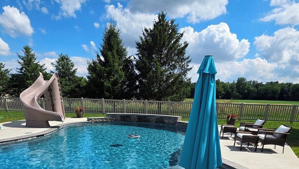 A swimming pool with a slide, a blue umbrella, and a deck with chairs. - LUXURY COUNTRY RETREAT ~ HEATED POOL, PICKLEBALL COURT, FIRE PIT & COUNTRY VIEWS (Wauseon)