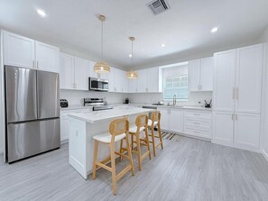 Uma cozinha moderna com armários brancos, eletrodomésticos de aço inoxidável, uma ilha central com banquetas, iluminação pendente e uma janela que proporciona luz natural.