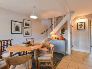 A dining area with a round table, wooden chairs, a staircase, and a kitchen area with a lamp. - Acorn Cottage South Cerney, 3 Double Bedrooms in the heart of the village (South Cerney)