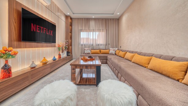A modern living room with a large flat-screen TV, a wooden coffee table, a beige sofa with orange cushions, and a fluffy white rug. - Luxury Design Apartment in New Residence with Two Pools, Close to All Amenities (Marrakesh)