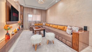 A modern living room with a large flat-screen TV, a sectional sofa, a wooden coffee table, and a decorative vase with flowers. - Luxury Design Apartment in New Residence with Two Pools, Close to All Amenities (Marrakesh)