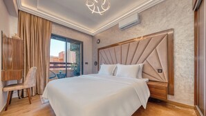 A modern bedroom with a large bed, a wooden nightstand, a chair, and a view of the outdoors. - Luxury Design Apartment in New Residence with Two Pools, Close to All Amenities (Marrakesh)
