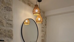A bathroom with a stone wall, a round mirror, a white sink, wooden countertop, and two hanging pendant lights. - Flexible & comfy guest room with mountain view (Montaillou)