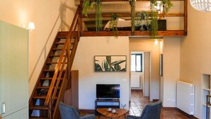 Ett vardagsrum i loftstil med en trätrappa, en platt-TV, två fåtöljer och hängande växter.