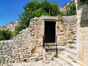 A stone staircase leading to a doorway with a metal gate, surrounded by greenery and a view of a hillside town.