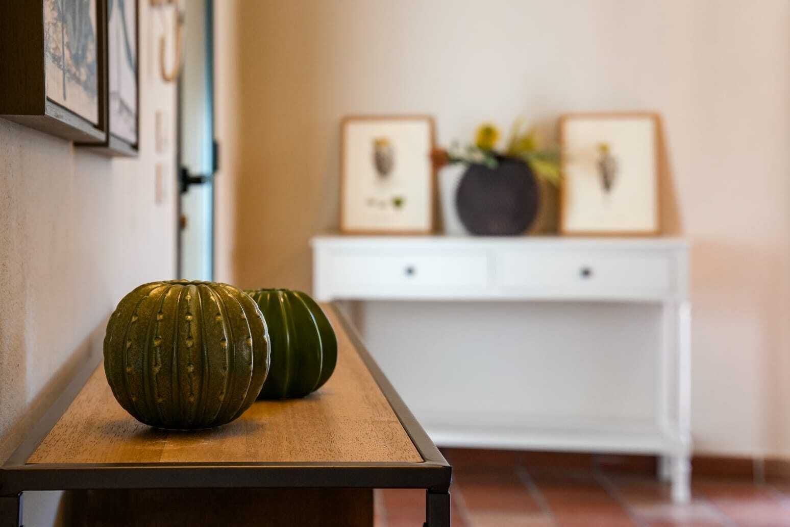 Un pasillo con un banco de madera, dos calabazas verdes, una consola blanca con marcos y un jarrón con flores.