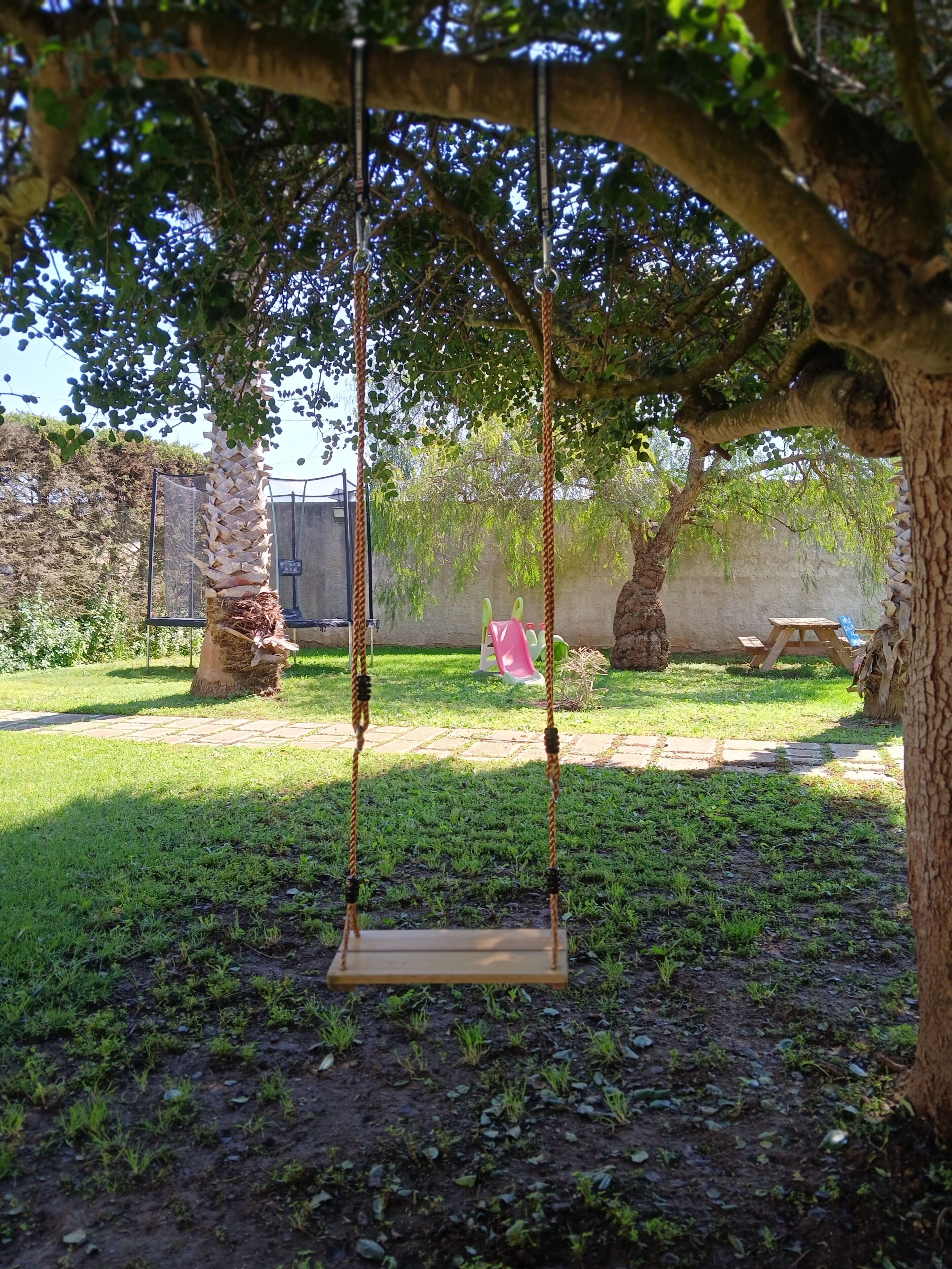 Une balançoire sous un arbre, dans un jardin avec un mur de pierres et une table de pique-nique en arrière-plan.