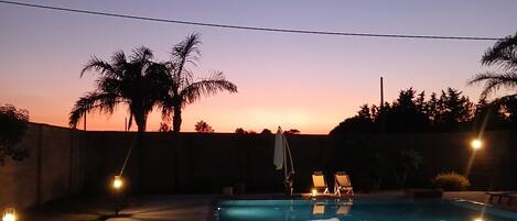 A swimming pool at dusk with a sunset in the background, surrounded by palm trees and a low wall.