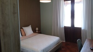 Een slaapkamer met een bed, nachtkastje, lamp, raam met gordijnen en een deur.