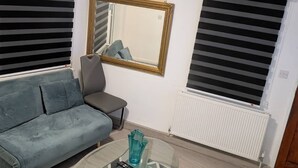 Ein Wohnzimmer mit einer Couch, einem Sessel, einem Glastisch, einem Spiegel und zwei Fenstern mit Jalousien.