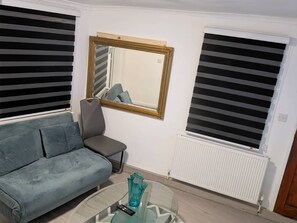 A living room with a sofa, a chair, a glass coffee table, a mirror, and two windows with blinds. - I bedroom Apartmwnt Goldbeater’s Grove (London)