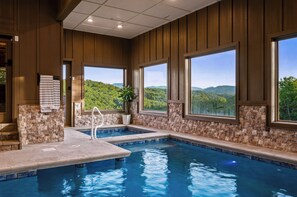 An indoor swimming pool with stone walls and large windows offering a view of a forested landscape. - Indoor Pool | Golf Sim | Pickleball | Sleeps 24 (Sevierville)