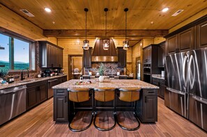 A kitchen with wooden ceiling, granite countertops, stainless steel appliances, and a large island with bar stools. - Indoor Pool | Golf Sim | Pickleball | Sleeps 20 (Sevierville)