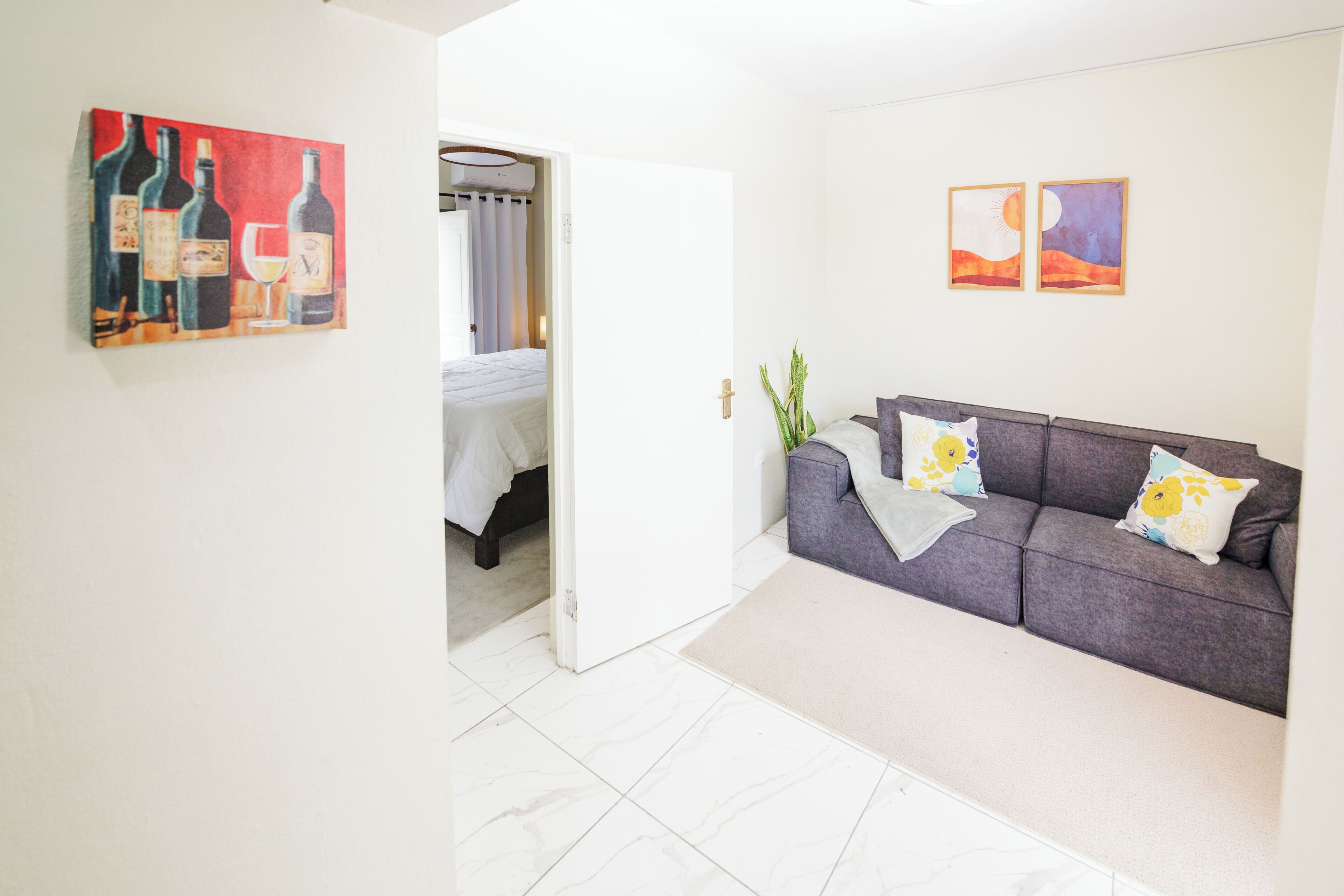 A room with a grey sofa, a bed with white bedding, a painting of wine bottles, and a door leading to another room.