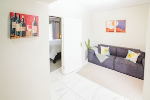 Ein Zimmer mit grauem Sofa, einem Bett mit weißer Bettwäsche, einem Gemälde mit Weinflaschen und einer Tür zu einem anderen Raum.