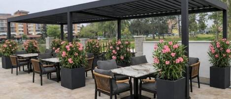 An outdoor patio area with tables and chairs, surrounded by potted plants with pink flowers, under a black pergola.