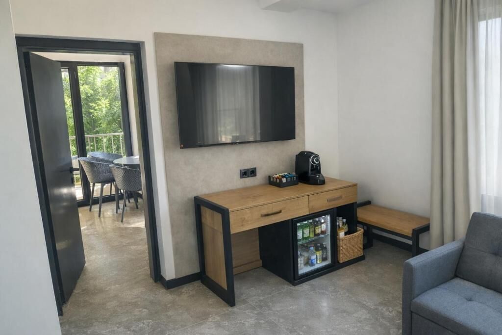 A modern hotel room with a flat-screen TV, a wooden desk with a coffee maker, a mini-fridge, and a seating area with a grey sofa.
