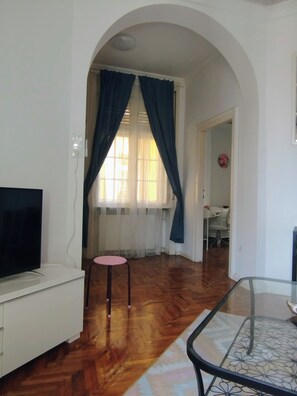Ein Zimmer mit Fernseher, einem kleinen pinkfarbenen Hocker, einem Glastisch und Holzboden.