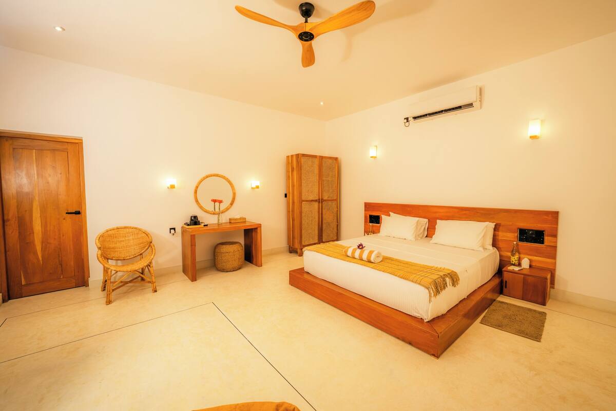 A bedroom with a wooden bed, a ceiling fan, a bedside table, a nightstand, a chair, a small table, and a mirror.