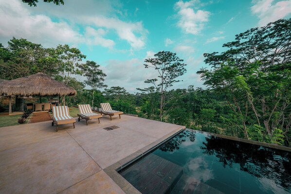 A pool area with lounge chairs, a thatched-roof structure, and lush greenery. - The 180 villa (Galle)