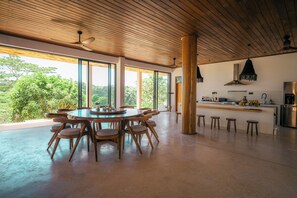 A spacious dining area with a round table and chairs, large windows with a view of greenery, a kitchen area with a bar, and a ceiling fan. - The 180 villa (Galle)