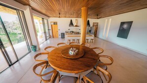 A modern dining area with a round wooden table surrounded by chairs, a kitchen in the background, and large windows offering a view of greenery. - The 180 villa (Galle)