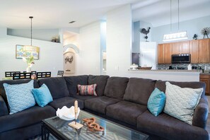 Ein Wohnzimmer mit einem Ecksofa, einem Glastisch und einer Küchenzeile mit Holzausbau und einer Mikrowelle.