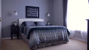 Une chambre à coucher avec un lit, deux tables de chevet, une lampe fixée au mur, une fenêtre avec des rideaux et une citation inspirante sur le mur.