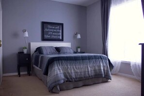 A bedroom with a bed, two bedside tables, a wall-mounted light, a window with curtains, and a motivational quote on the wall.