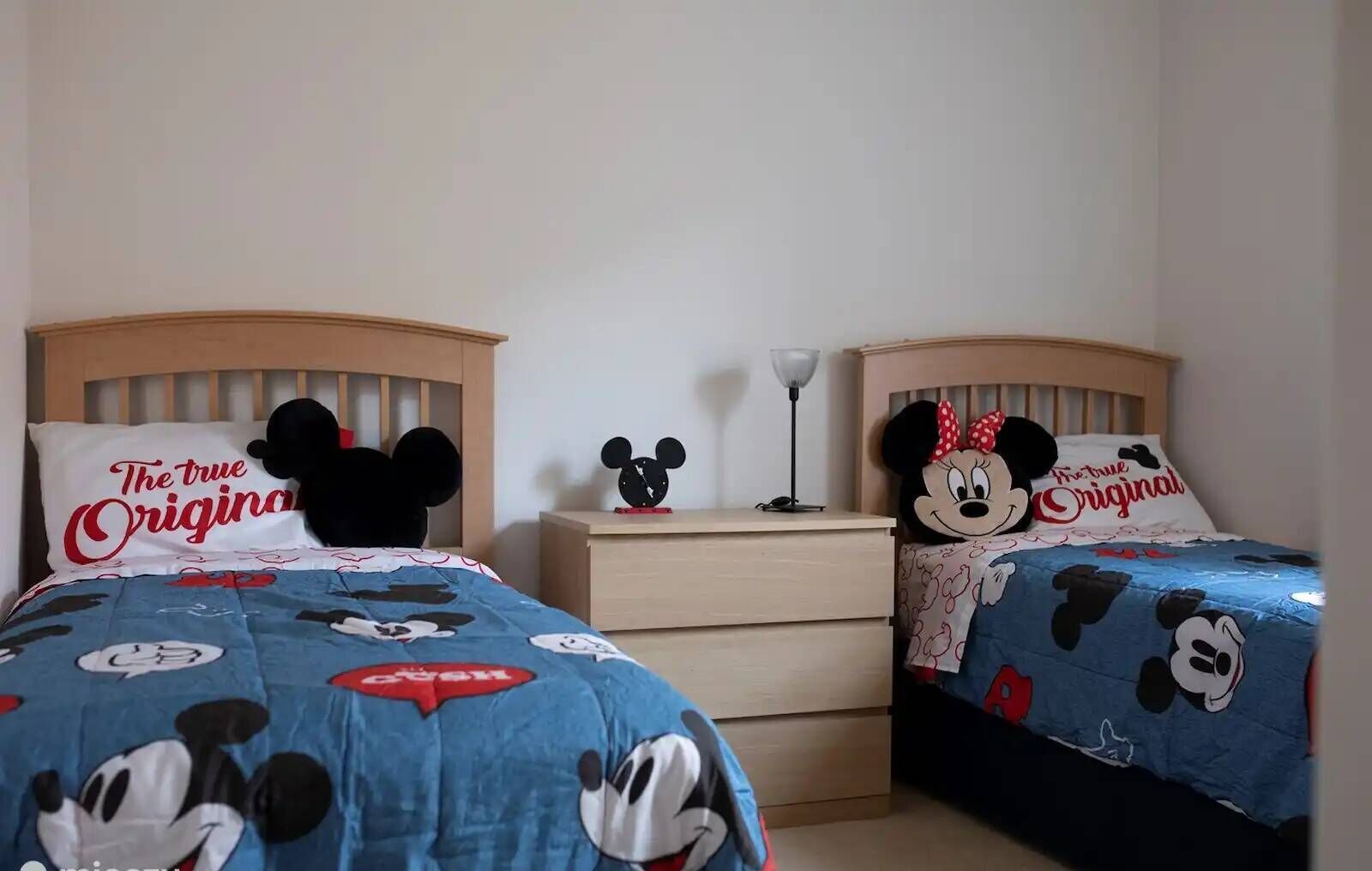 Deux lits jumeaux avec une literie aux motifs de Mickey Mouse, une table de chevet avec une lampe et un oreiller orné de l’inscription « The true Originals ».