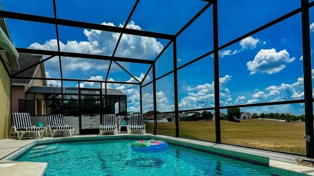 Une piscine à l’eau d’un bleu cristallin, entourée de grandes fenêtres qui offrent une vue sur un champ verdoyant et un ciel dégagé.