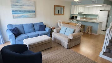 A living room with a blue sofa, a beige armchair, and a coffee table. There is a kitchen in the background with white cabinets and a microwave.